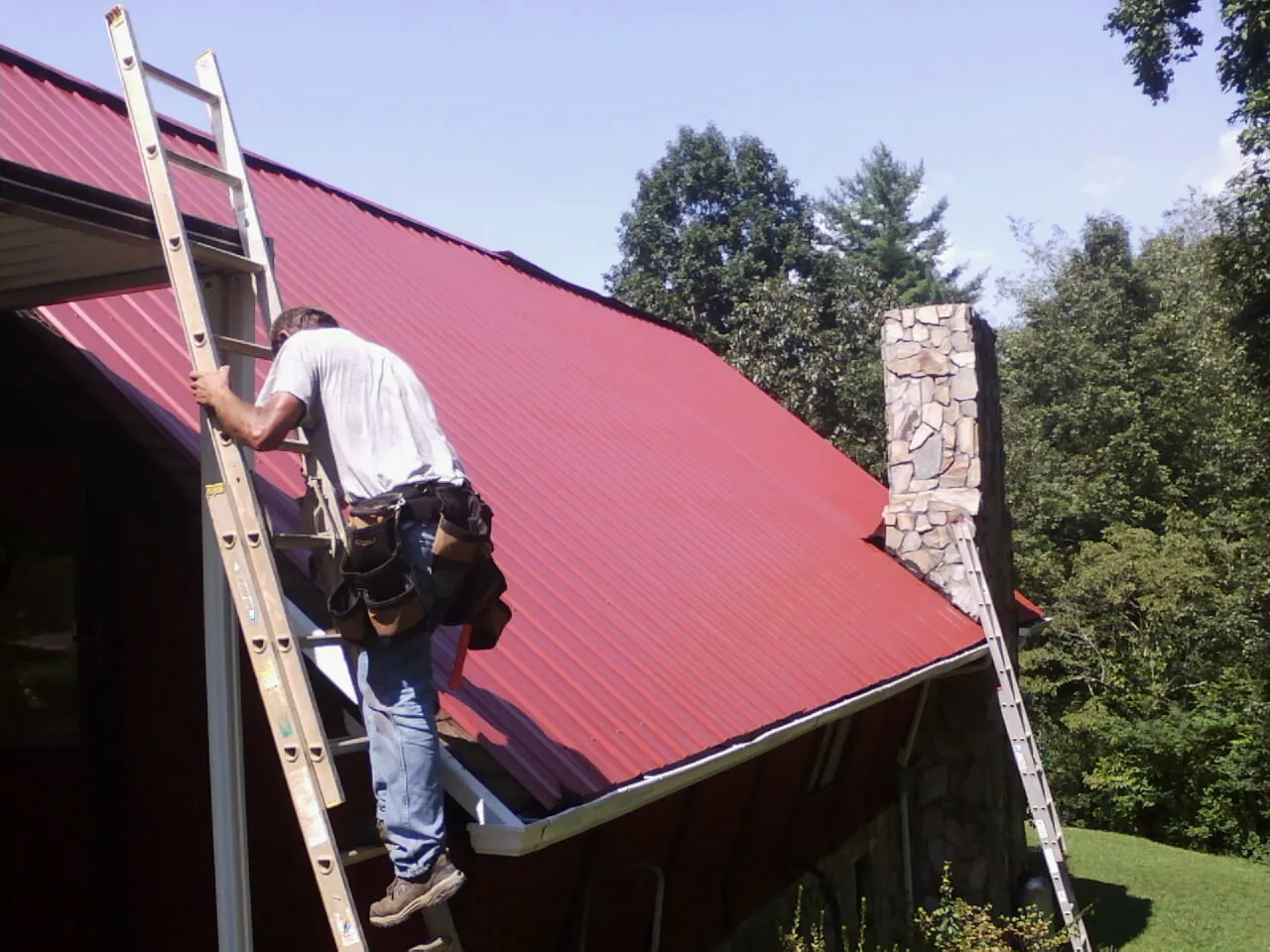 Roofer on a ladder beside a red metal roof and stone chimney — Soffit & Fascia Repair by Roofing Brothers in Saint Clair Shores